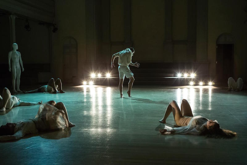 Dancers lay on their back as another male dancer in white strikes a standing pose with his fingers splayed